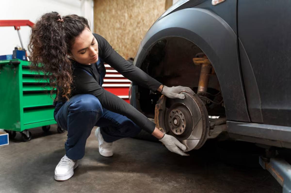 A person in gloves kneels beside a black car, working on the brake rotor in a garage filled with tools and equipment.