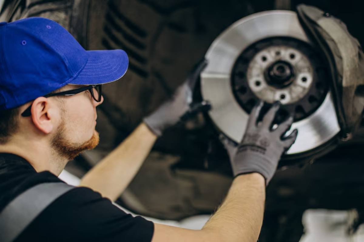 A mechanic in a blue cap and gloves works on a vehicle’s brake system, holding a disc brake in a garage setting.