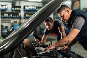 Two mechanics work on a car engine in a garage, using a laptop and diagnostic tools to assess the vehicle.