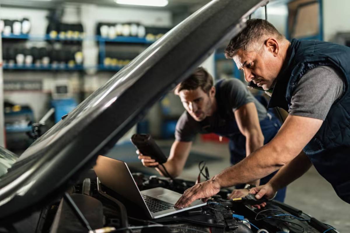 Two mechanics work on a car engine in a garage, using a laptop and diagnostic tools to assess the vehicle.