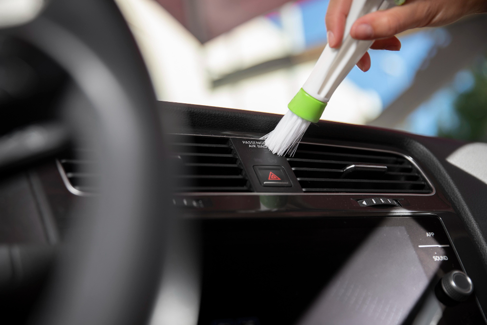 A hand uses a small brush to clean the air vent and controls on a car's dashboard, with a focus on tidiness and maintenance.