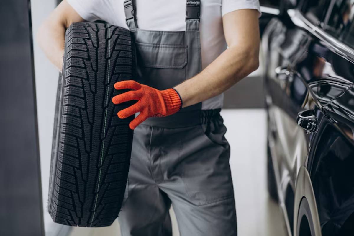A mechanic in gray overalls and an orange glove holds a new tire, preparing to install it on a black vehicle in a garage.