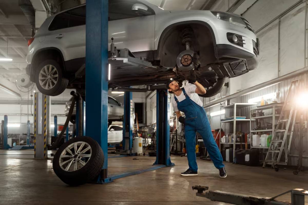 A mechanic in blue overalls inspects the undercarriage of a lifted silver SUV in a well-equipped auto repair shop. Tires and tools nearby.