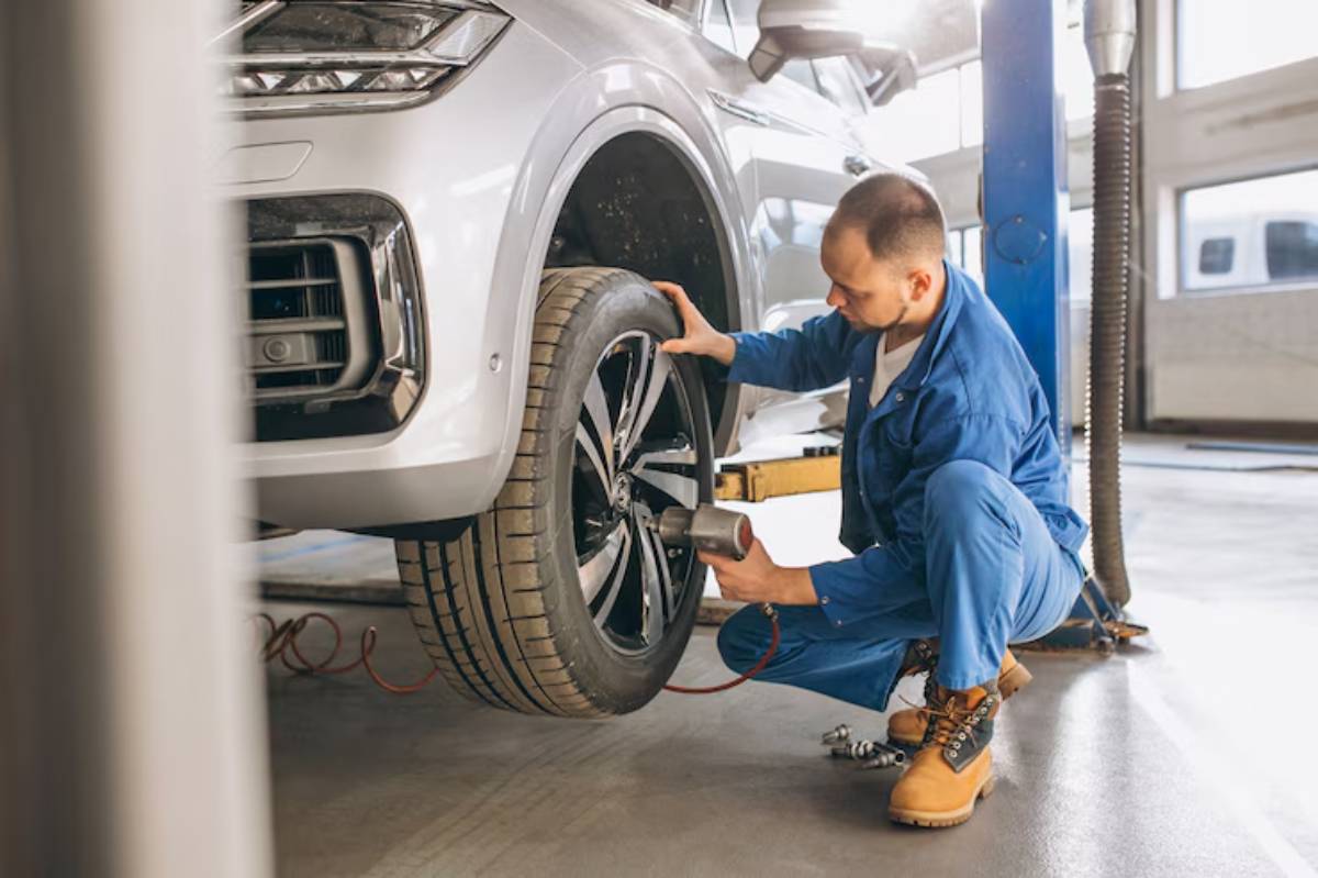A mechanic in blue overalls uses a power tool to attach a black wheel to a silver SUV in a bright garage setting.