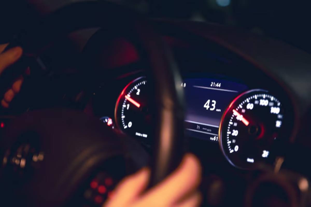Close-up of hands holding a steering wheel, with a dashboard showing a speed of 43 mph and a time of 21:44, in low light.