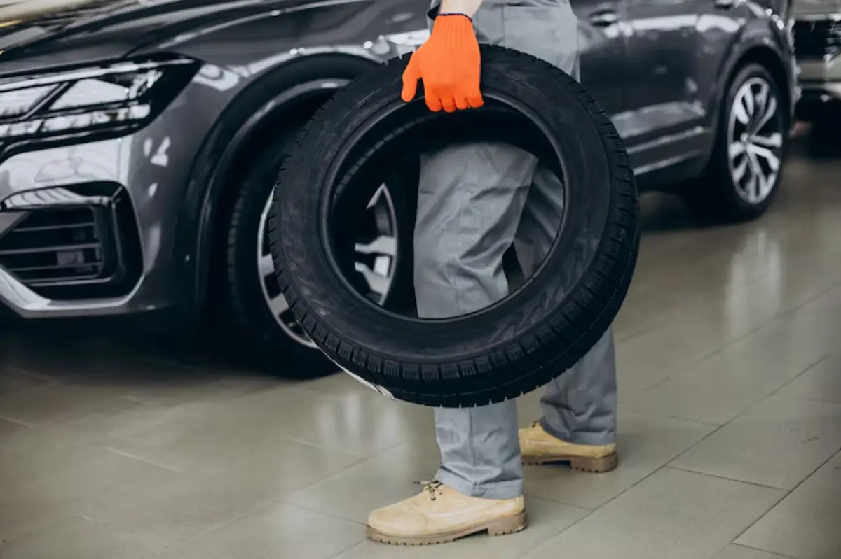 A person in gray overalls and orange gloves holds a tire near a shiny gray car in a well-lit automotive showroom.