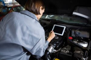 A female mechanic examines a car engine, using a tablet for diagnostics in a well-lit workshop.