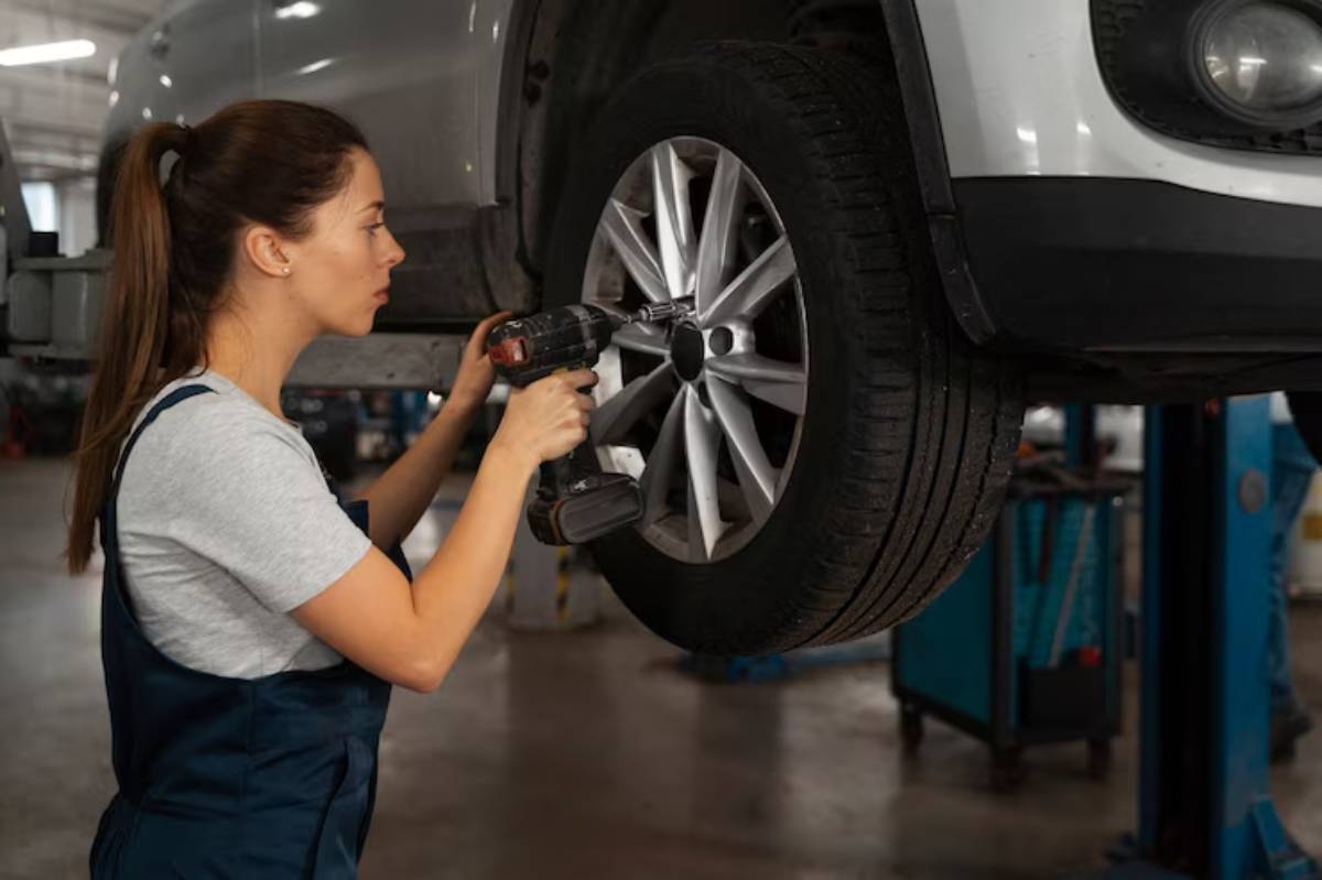 A woman in a gray shirt and blue overalls uses a power tool to work on a vehicle's wheel in a garage setting.