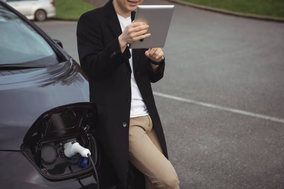 A man in a black coat smiles while using a tablet, standing next to an electric vehicle charging station.