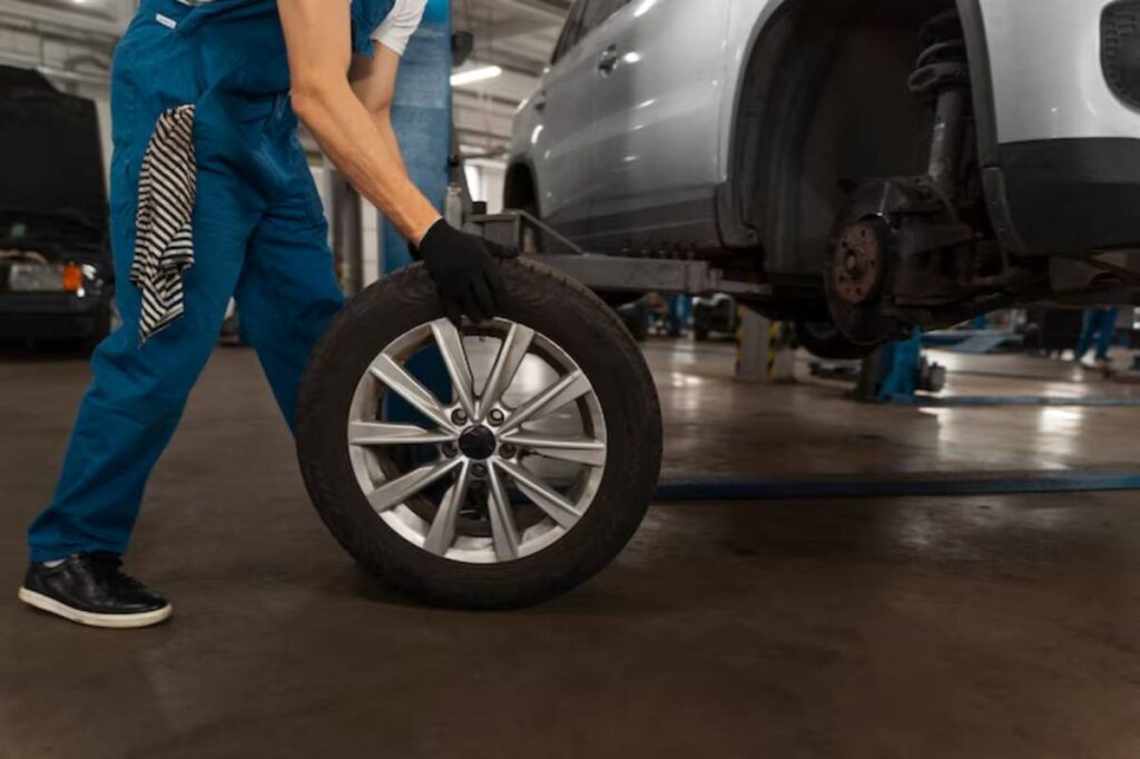 A mechanic lifts a car tire while working on a silver vehicle in a well-lit auto repair shop.