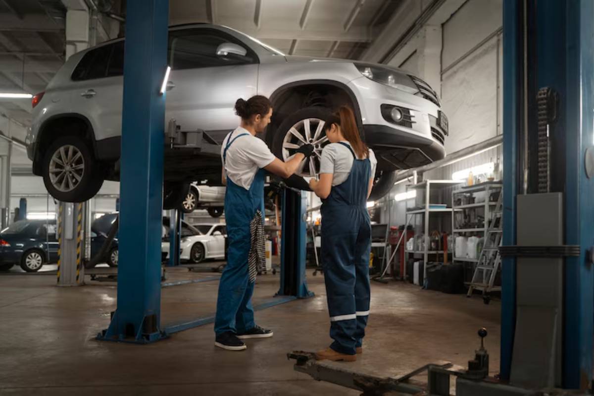 Two mechanics in blue overalls working on a silver car elevated on a lift in a well-lit garage, surrounded by other vehicles.