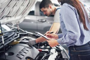 A woman holding a clipboard observes a mechanic inspecting a car engine in a garage. Various tools and parts are visible.