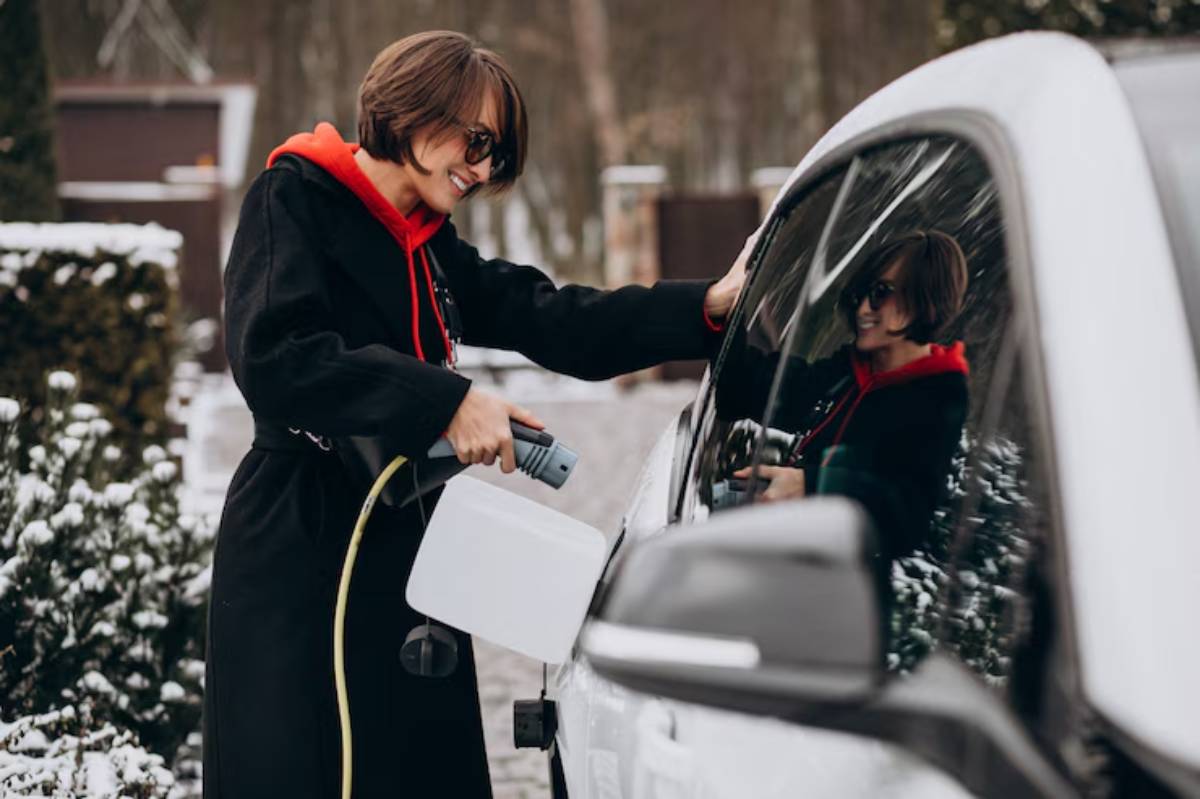 A person in a black coat and red hoodie is filling a car with liquid using a funnel in a snowy outdoor setting.