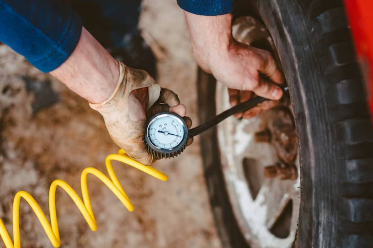 A person wearing gloves checks tire pressure with a gauge connected to a tire, with a yellow air hose coiled nearby.