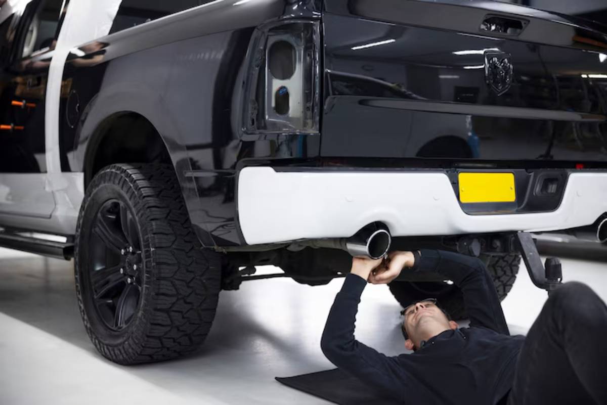 A person works underneath a black and white truck, inspecting the exhaust system in a well-lit garage setting.