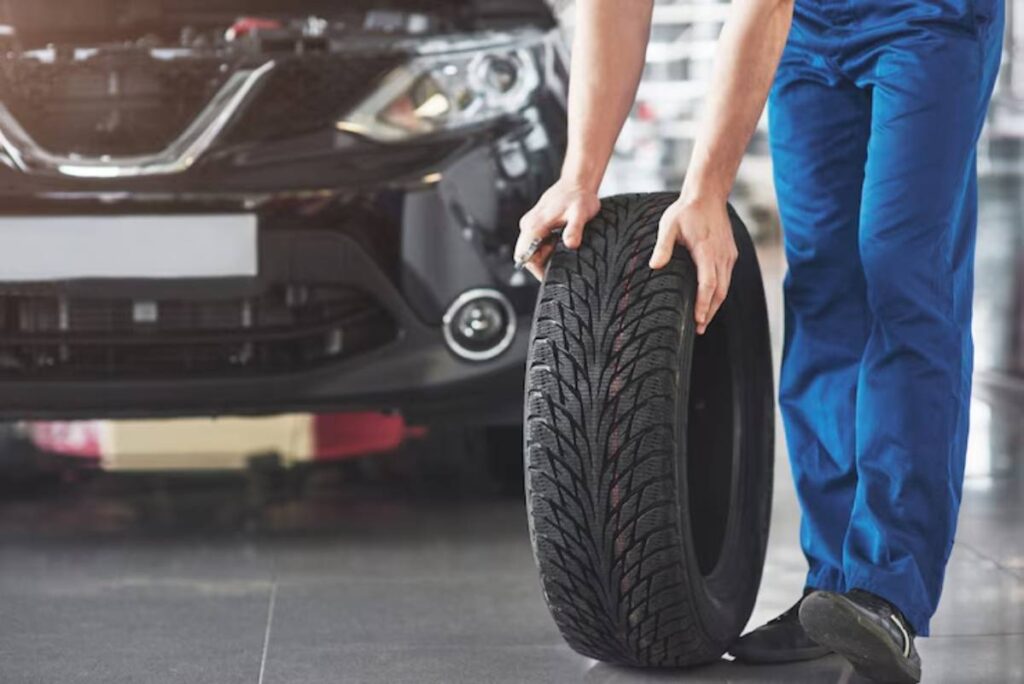 A mechanic in blue overalls holds a new tire in front of a black car in a garage, preparing for a tire change or inspection.