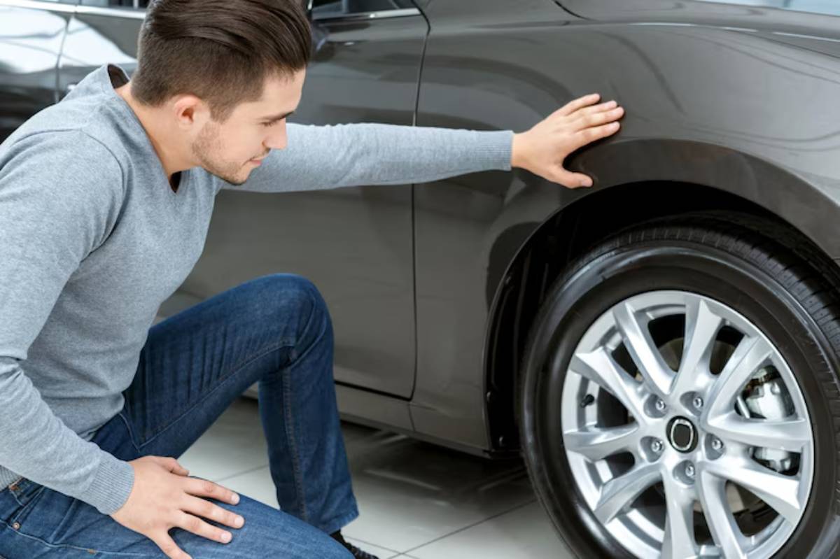 A man inspects a gray car, kneeling to touch the side panel while examining the vehicle's details in a showroom.
