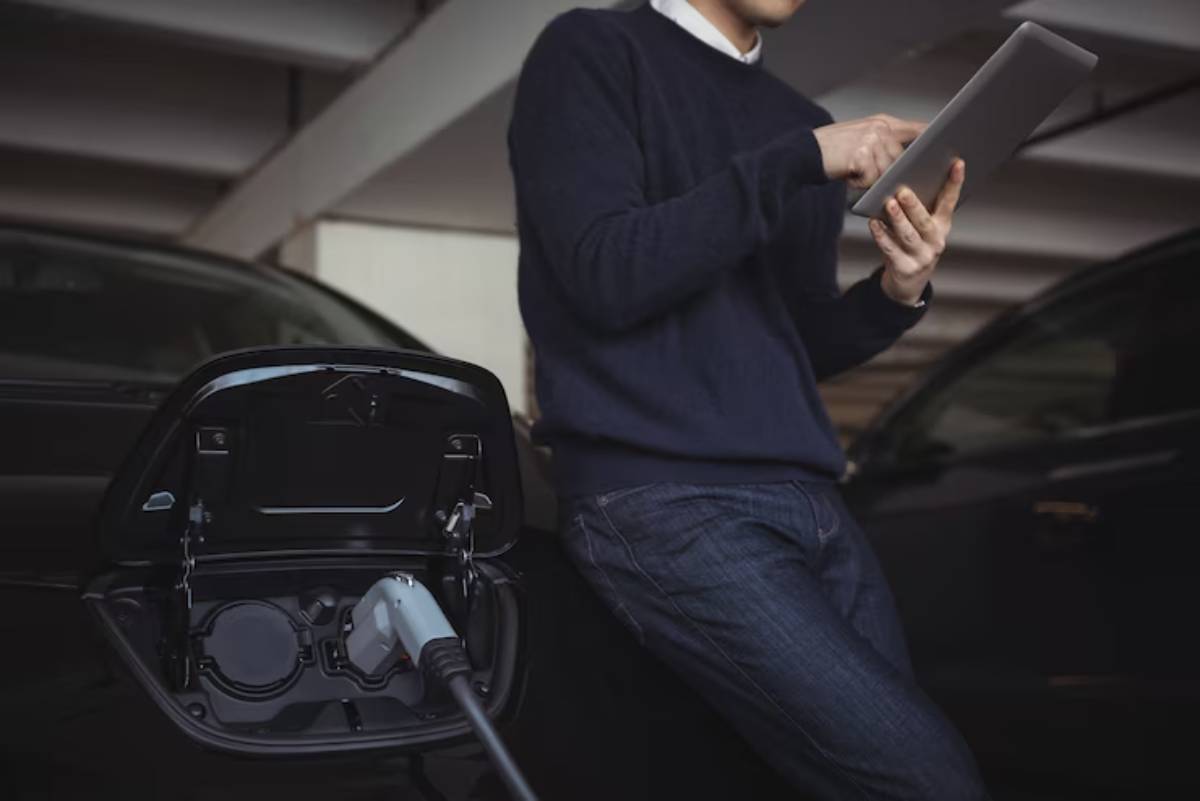 A man in a sweater uses a tablet while standing next to an electric vehicle charging in a garage.