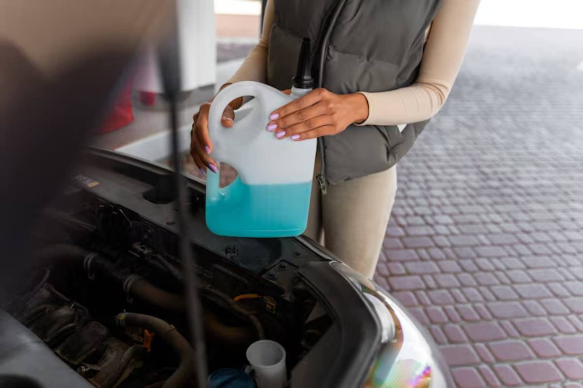 A person in a gray vest pours blue windshield washer fluid from a jug into the open hood of a car.