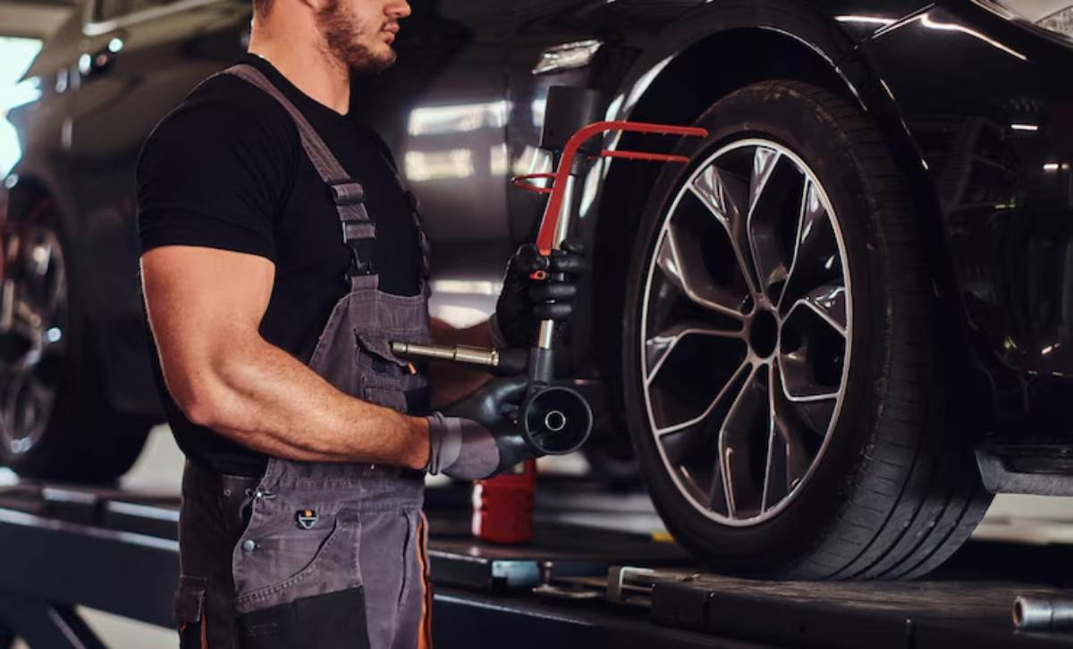A mechanic holds tools, preparing to work on a vehicle's wheel in a well-lit garage. The car is raised on a service platform.