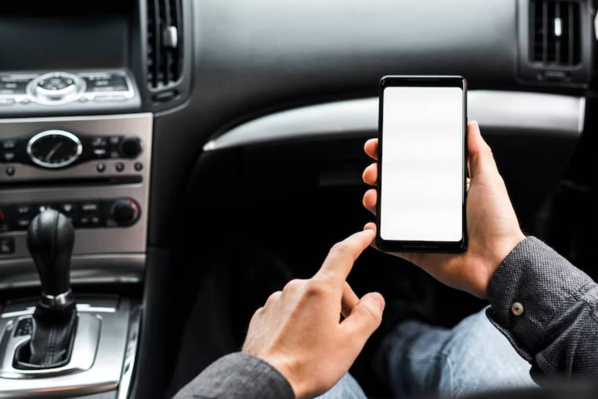 A person holds a smartphone displaying a blank screen while seated in a car, with gear shift and dashboard controls visible.