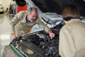 Two mechanics in beige overalls are examining an open car hood, focused on the engine components. Tools are scattered nearby.