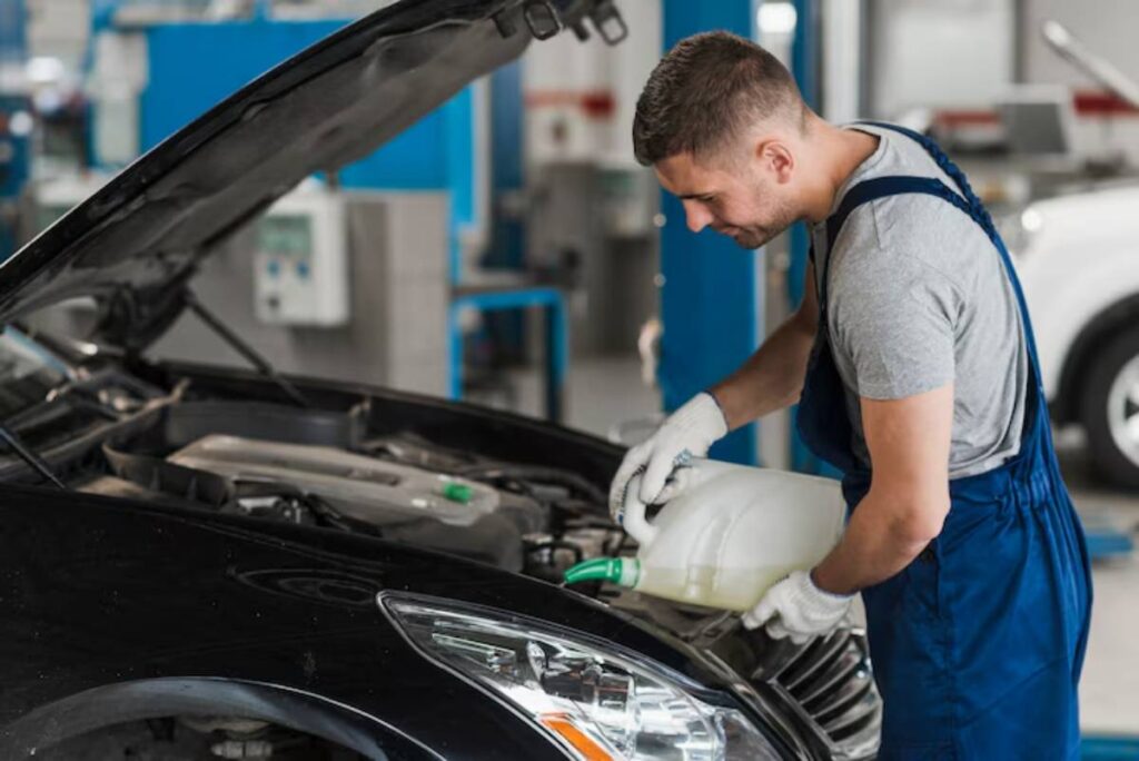 A mechanic in blue overalls pours fluid into the engine compartment of a black car in a workshop. Tools and equipment are visible in the background.