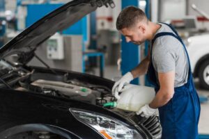 A mechanic in blue overalls pours fluid into the engine compartment of a black car in a workshop. Tools and equipment are visible in the background.
