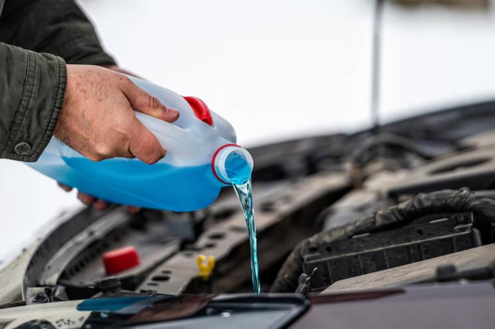 A person pours blue windshield fluid from a jug into a car's engine compartment, surrounded by a snowy background.