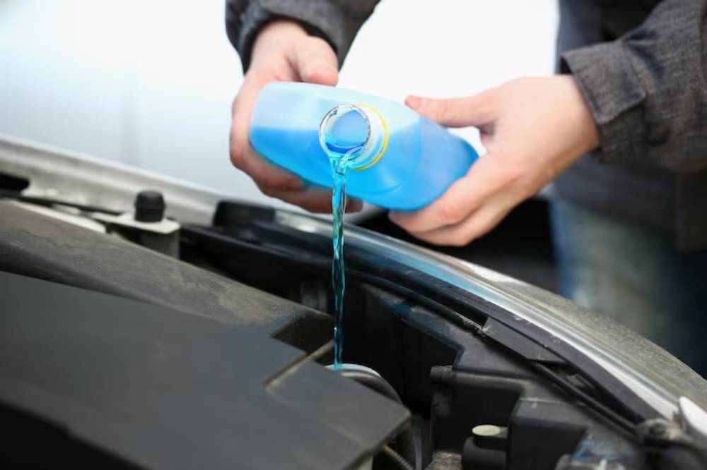 Person pouring blue liquid from a container into a car engine, performing vehicle maintenance under the hood.