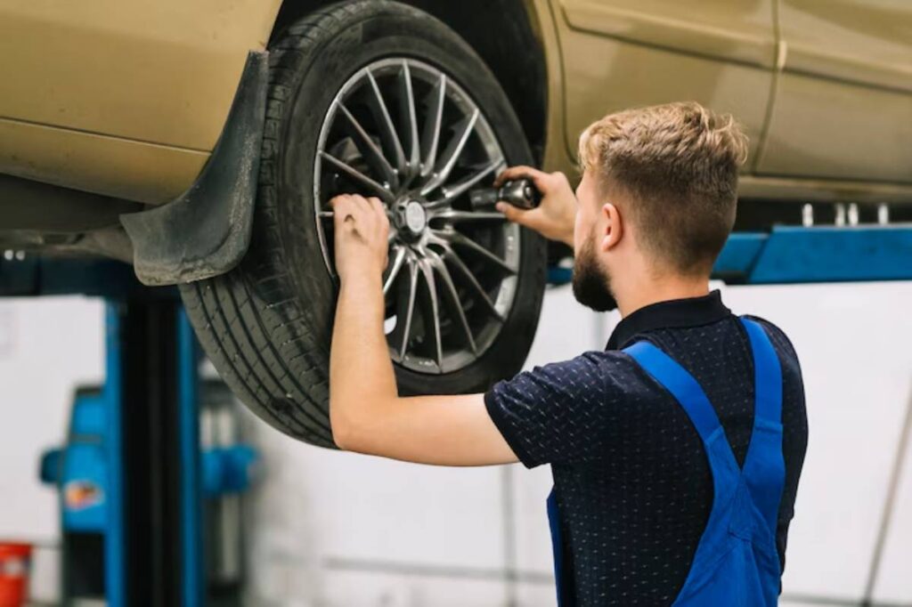 A mechanic in blue overalls uses a wrench to adjust a wheel on a lifted gold car in a garage.
