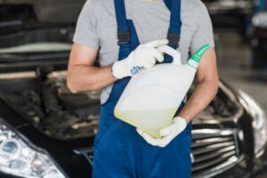 A mechanic in gloves holds a gallon jug of yellow fluid, standing in front of a car's open hood in a garage setting.