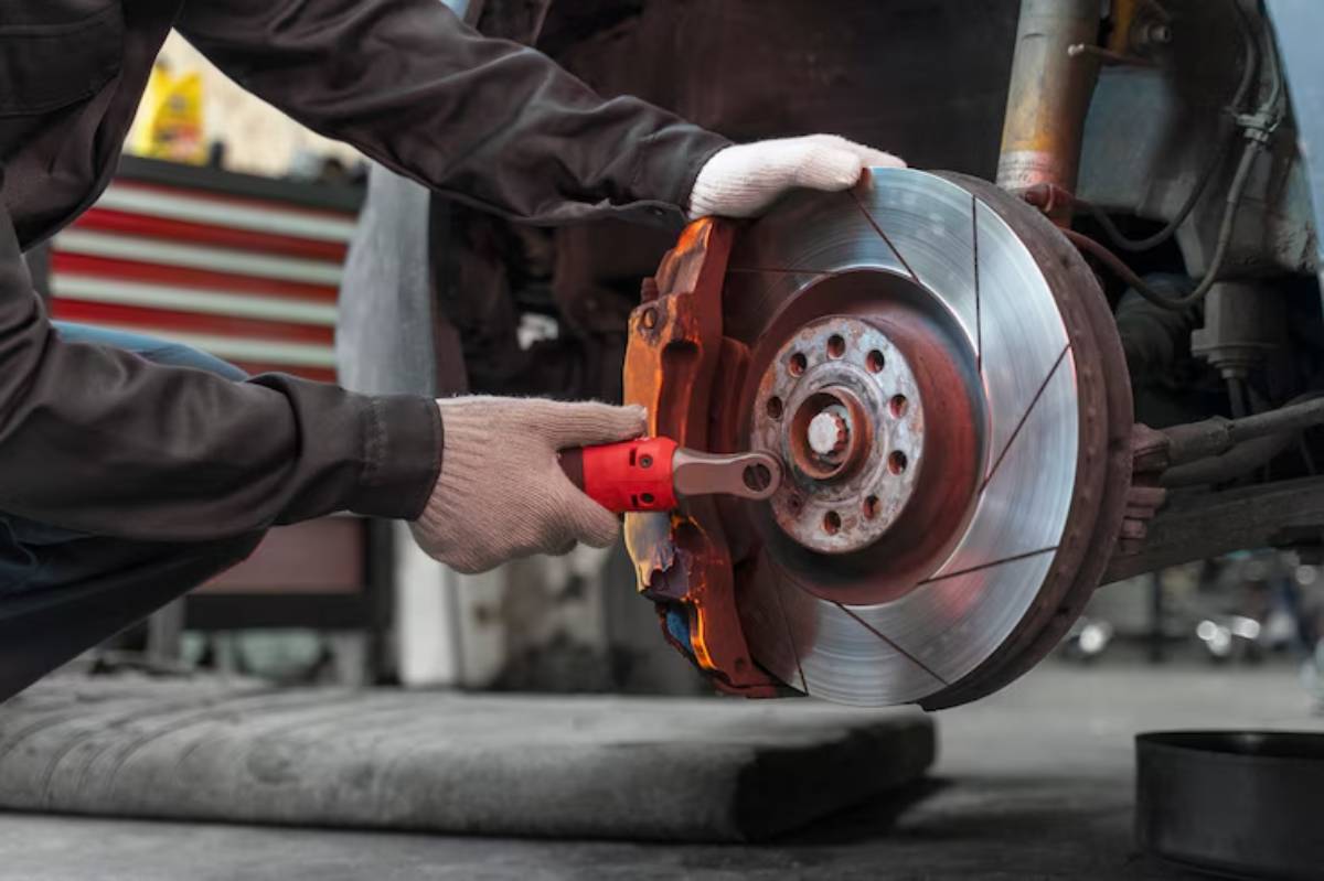 A mechanic inspects and works on a car's brake system using a ratchet tool, wearing gloves in a workshop setting.