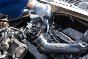 A mechanic in gloves uses a wrench to work on the engine of a vehicle under the hood. Various engine components are visible.