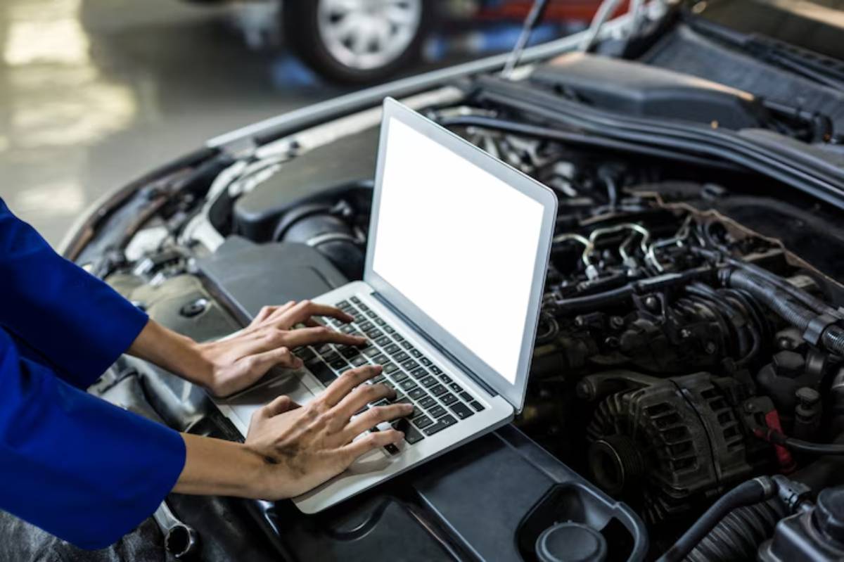 A mechanic in a blue uniform types on a laptop placed on a car engine, surrounded by vehicle parts in a workshop.