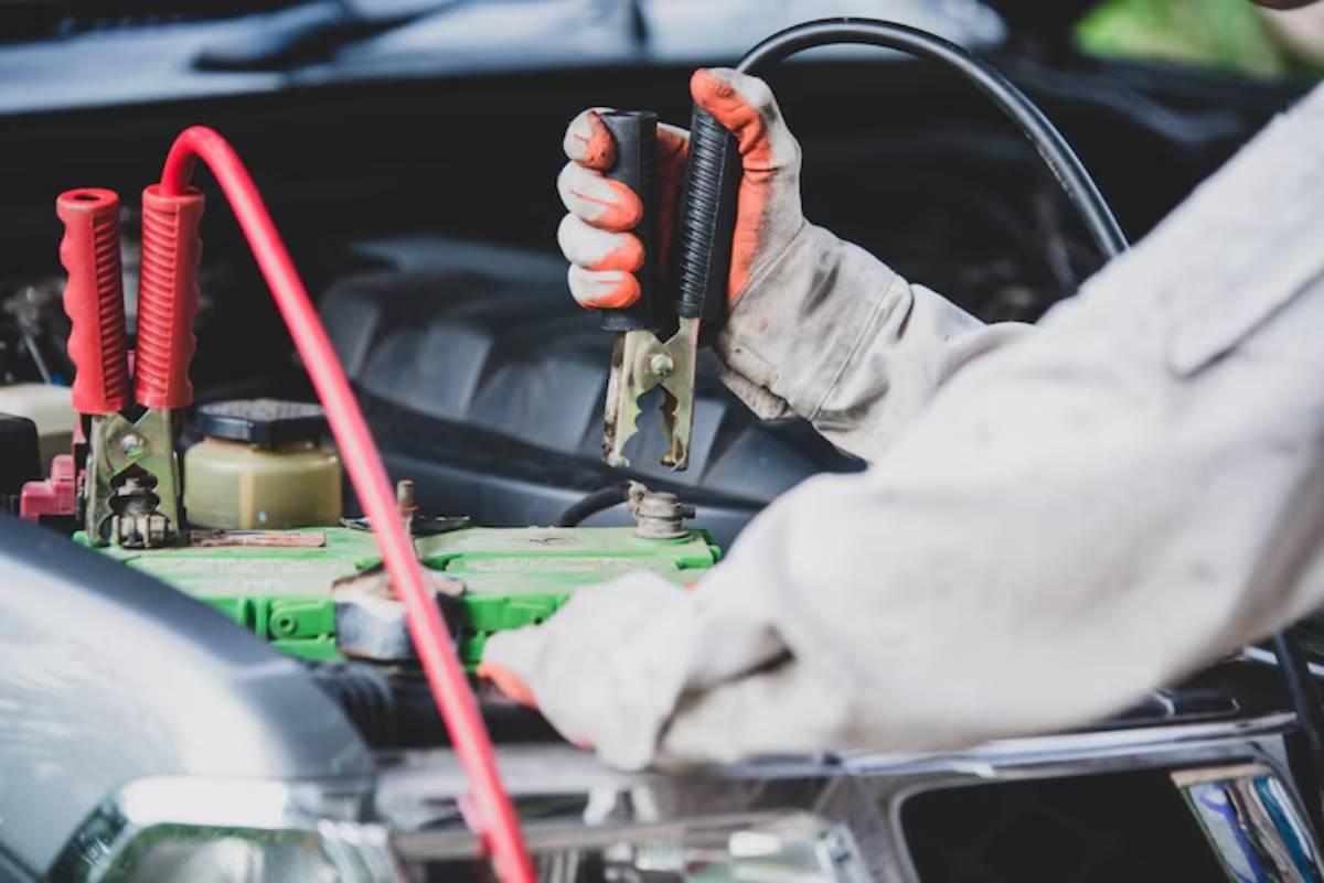 A mechanic in gloves holds a jumper cable clamp, preparing to connect it to a green car battery under a vehicle's hood.