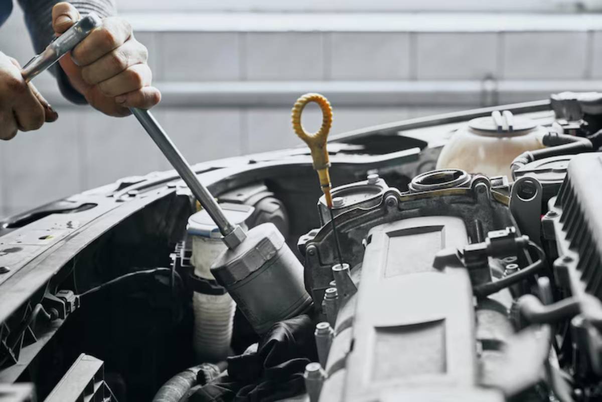 A mechanic uses a wrench to work on a car engine, with an oil dipstick visible in the foreground and various engine components around.