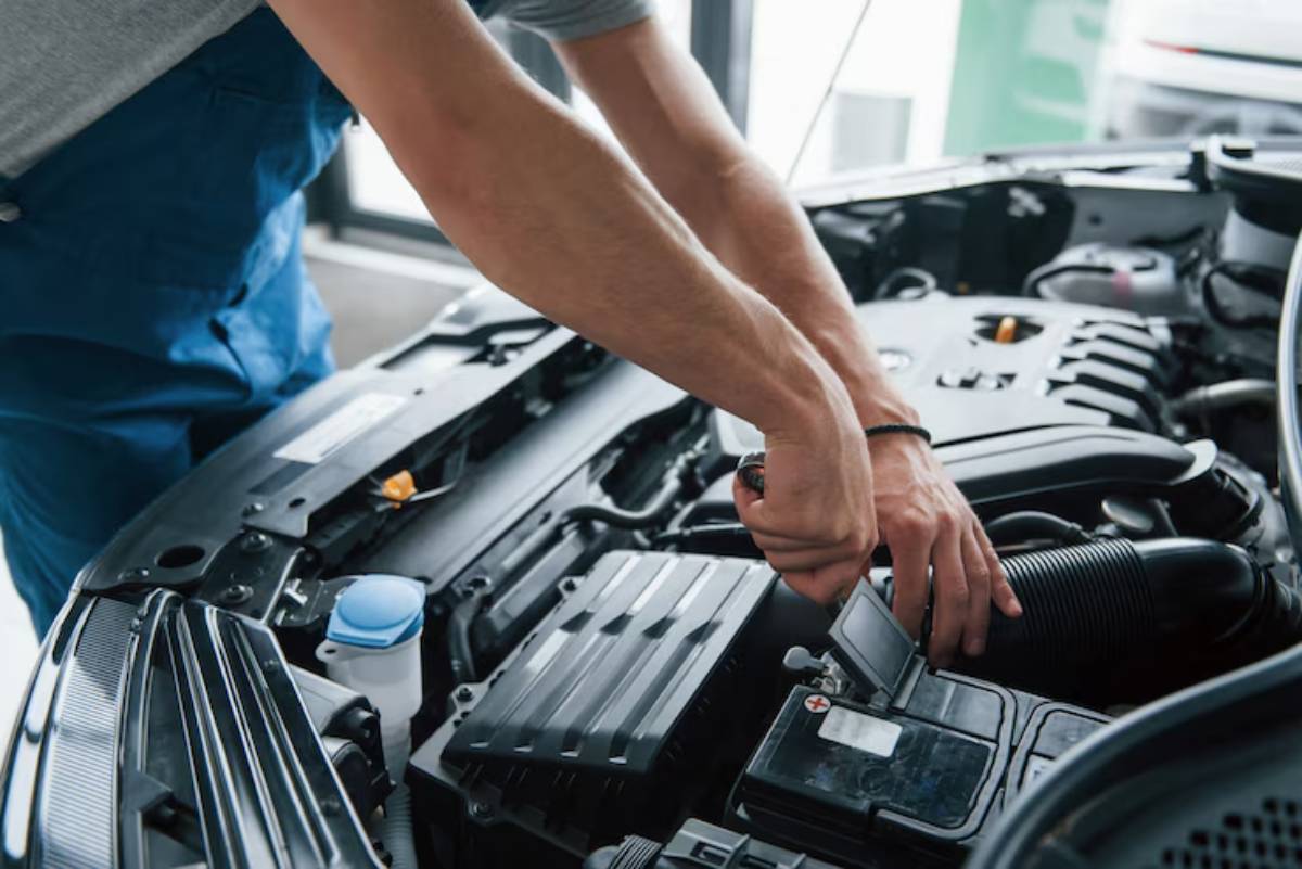 A mechanic in a gray shirt works on a car engine, adjusting components under the hood of a vehicle.