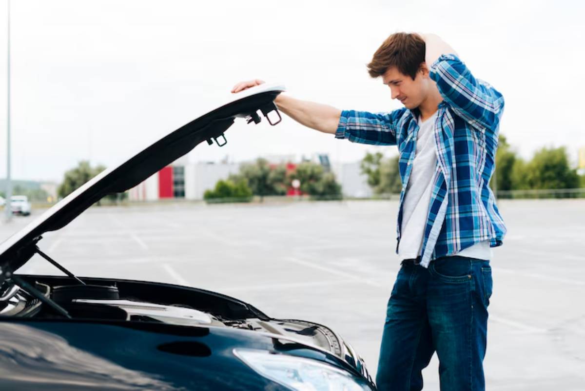 A person stands beside a black car with its hood raised, looking perplexed in an empty parking lot under a cloudy sky.