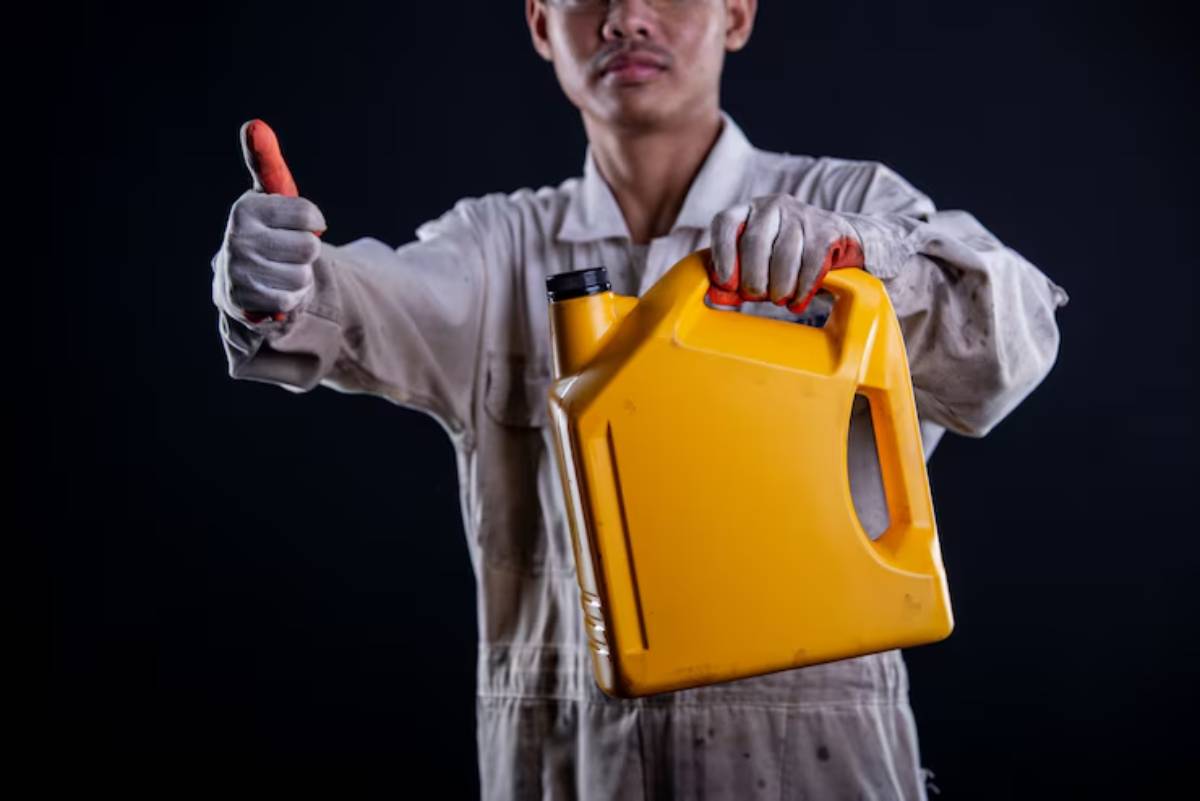 A person in a dirty work uniform holds a yellow container and gives a thumbs-up, suggesting satisfaction or approval.