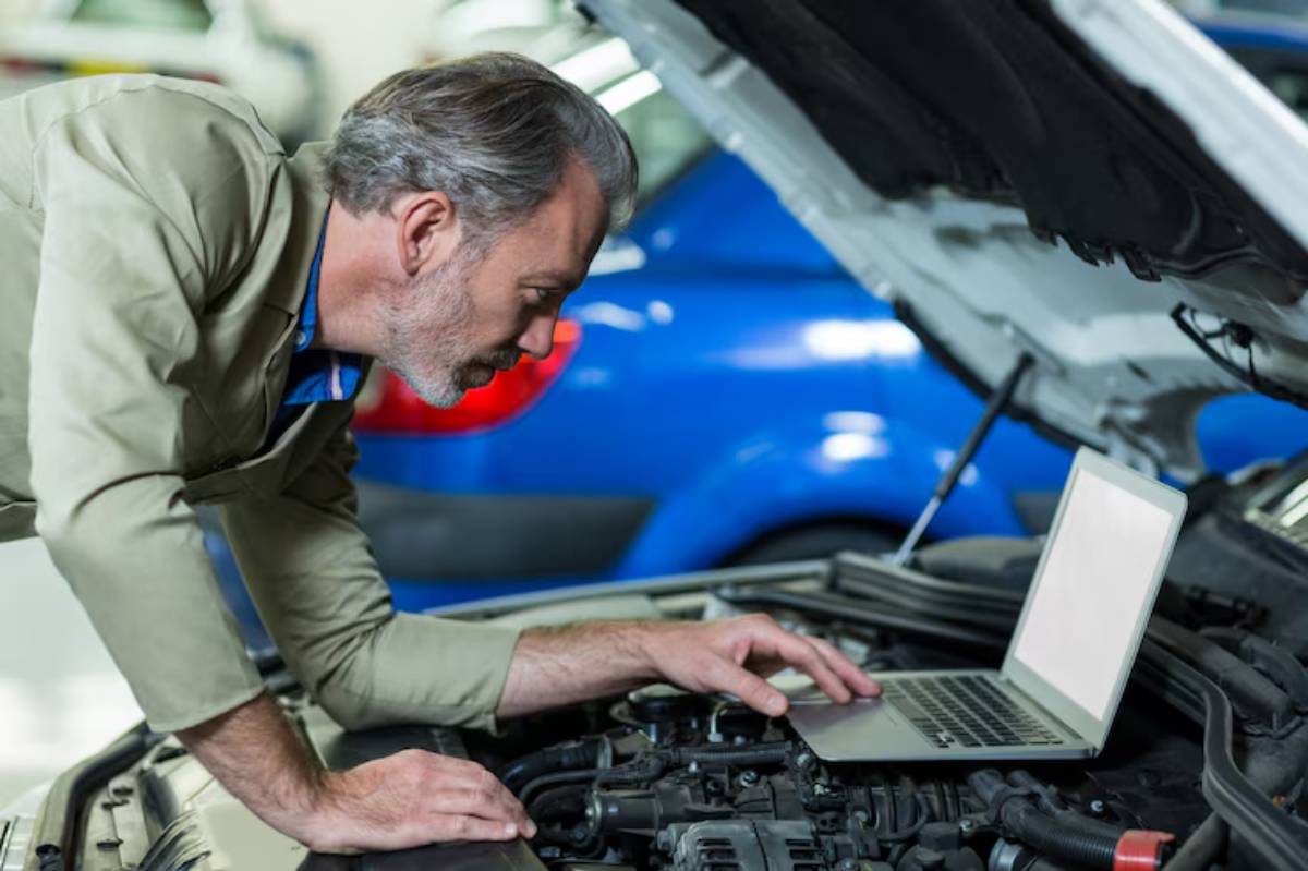 A mechanic in a beige jacket leans over an open car hood, using a laptop to diagnose or troubleshoot vehicle issues.