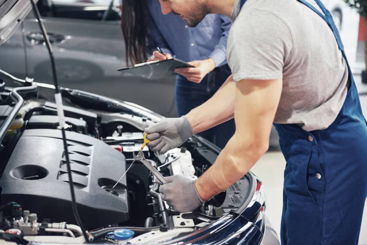 A mechanic in gray gloves uses a wrench to work on a car engine, while a woman observes with a clipboard.
