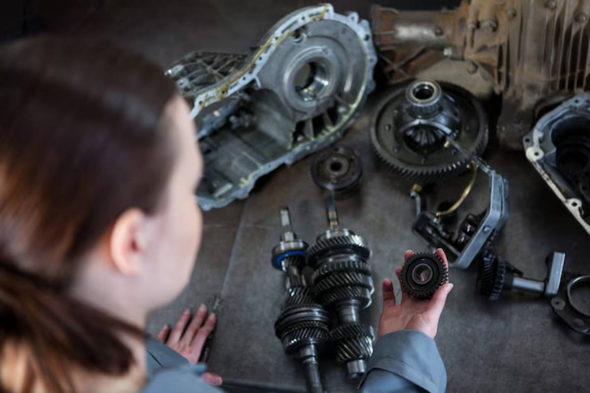 A person inspects various mechanical parts, including gears and an engine casing, on a work surface in a workshop setting.