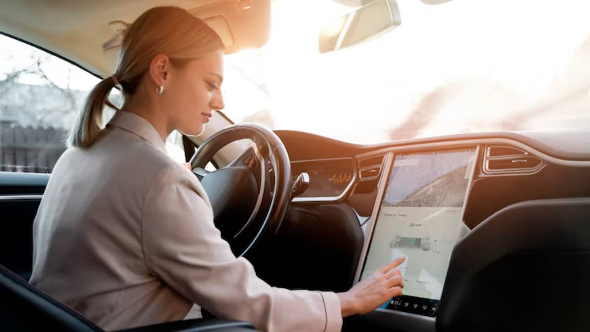 A woman in a car interacts with a large touchscreen display showing navigation details, with sunlight pouring in through the window.