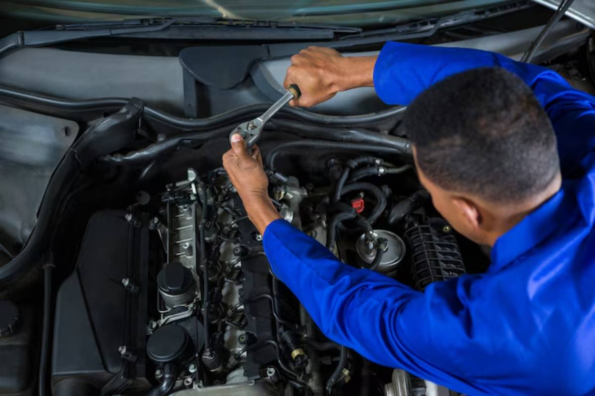 A mechanic in a blue uniform uses a wrench to service a car engine, focusing on the intricate components within the engine bay.