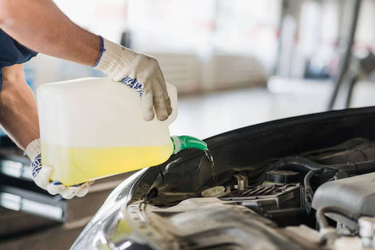 A mechanic pours yellow automotive fluid from a container into a car's engine compartment, wearing gloves for protection.