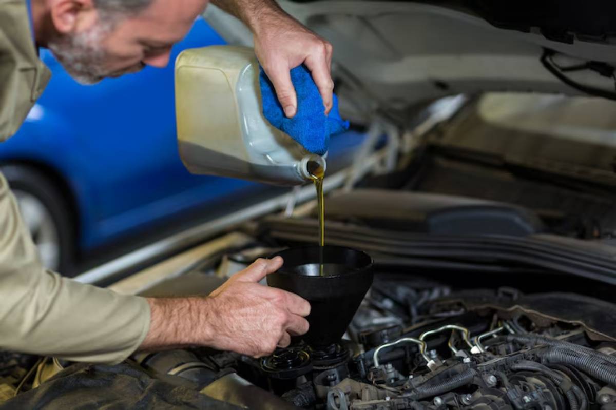 A person pours engine oil into a funnel while working on a car's engine, surrounded by tools and car parts in a garage.