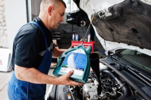A mechanic in a blue uniform pours oil from a green container into an engine using a funnel, surrounded by tools and equipment.